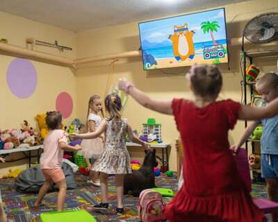 Чотири молоді дівчинки стрибають, дивлячись по телевізору, як мультяшний персонаж тренується у підвалі, повному м'яких іграшок.  